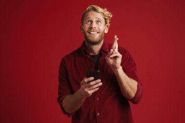 Beyaz sakallı adam parmaklarını çapraz yapıp kırmızı duvarda izole edilmiş bir cep telefonu kullanıyor.