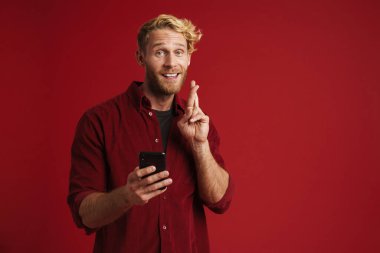 Beyaz sakallı adam parmaklarını çapraz yapıp kırmızı duvarda izole edilmiş bir cep telefonu kullanıyor.