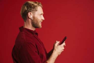 Sarışın adam kulaklıklı müzik dinliyor ve kırmızı duvardan izole edilmiş bir cep telefonu kullanıyor.