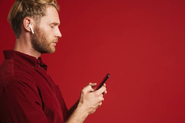 Sarışın adam kulaklıklı müzik dinliyor ve kırmızı duvardan izole edilmiş bir cep telefonu kullanıyor.