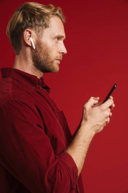 Sarışın adam kulaklıklı müzik dinliyor ve kırmızı duvardan izole edilmiş bir cep telefonu kullanıyor.