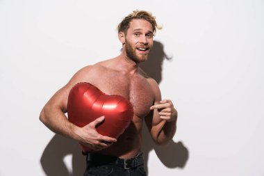 Beyaz arka planda izole edilmiş kalp balonunu işaret ederken gülümseyen gömleksiz sarışın adam.