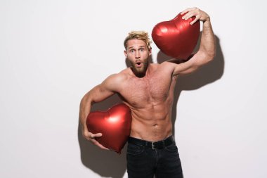 Beyaz duvarın üzerinden izole edilmiş şişme kalplerle poz veren gömleksiz bir adam şaşkınlığını dile getiriyor.