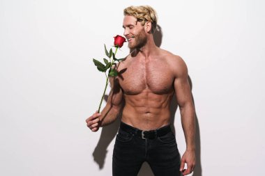 Beyaz arka planda kırmızı gülle poz verirken gülümseyen gömleksiz sarışın adam.