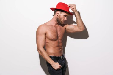 Beyaz gömleksiz adam yana bakıyor ve beyaz duvarın üzerinde kırmızı şapkayla poz veriyor.