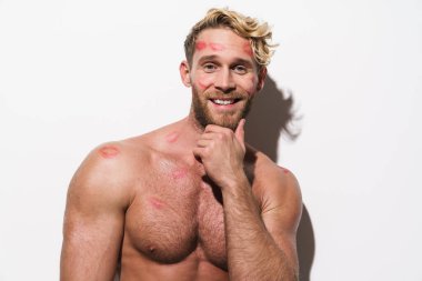 Beyaz tişörtsüz, ruj izi olan beyaz adam gülümseyen ve beyaz duvarın üzerinden izole edilmiş kameraya bakan.