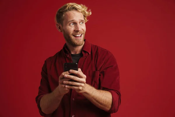 White bearded man wearing shirt laughing and using cellphone isolated over red wall