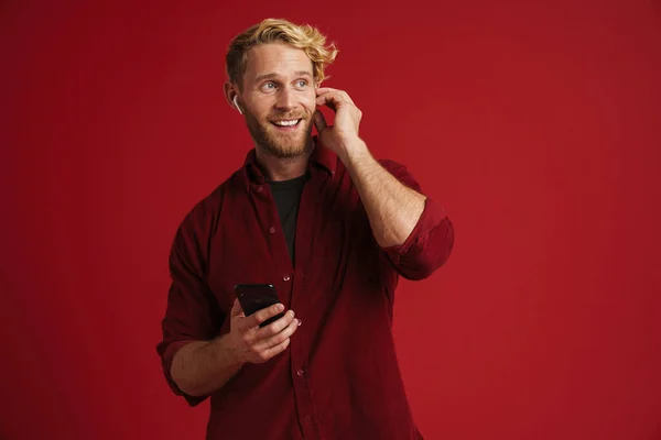 Blonde Man Listening Music Earphones Using Cellphone Isolated Red Wall — Stockfoto