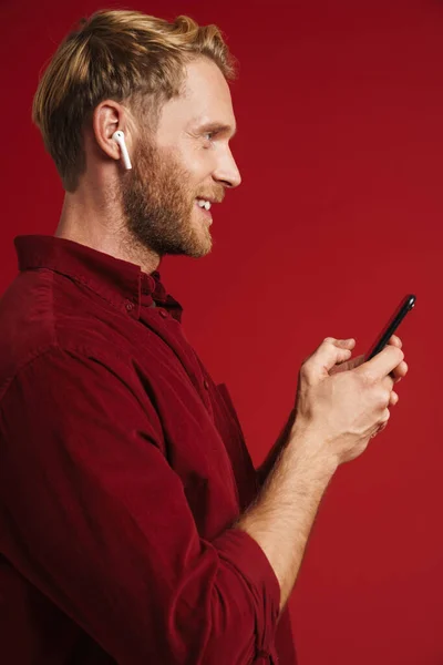 Blonde Man Listening Music Earphones Using Cellphone Isolated Red Wall — стоковое фото