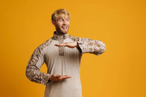 White military man wearing uniform smiling and holding copyspace isolated over yellow wall