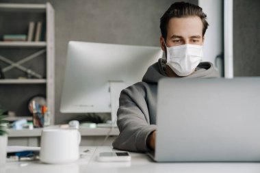 Young white man in protective mask working with laptop at office