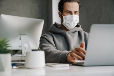 Young white man in protective mask working with laptop at office