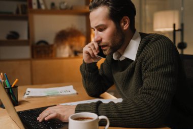 Ofiste dizüstü bilgisayarla çalışırken cep telefonuyla konuşan genç kıllı adam.