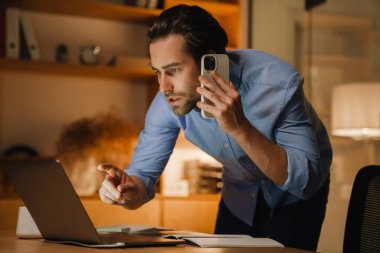 Ofiste dizüstü bilgisayarla çalışırken cep telefonuyla konuşan genç kıllı adam.