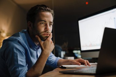 Genç adam ofiste otururken bilgisayar ve dizüstü bilgisayarla çalışıyor.