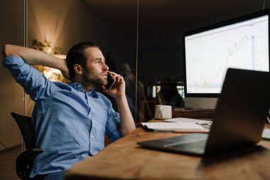 Ofiste dizüstü bilgisayarla çalışırken cep telefonuyla konuşan genç kıllı adam.