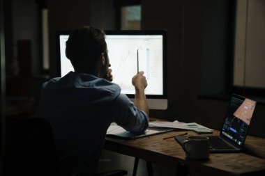 Genç adam ofiste otururken bilgisayar ve dizüstü bilgisayarla çalışıyor.