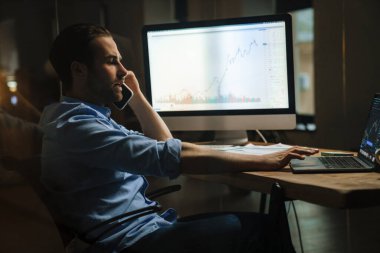 Ofiste dizüstü bilgisayarla çalışırken cep telefonuyla konuşan genç kıllı adam.