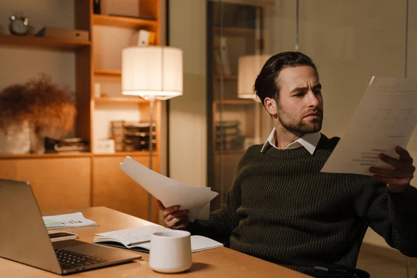 Young puzzled man working with laptop and papers in office