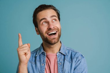 Young excited man wearing shirt smiling and pointing finger upward isolated over blue background