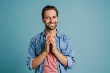 Genç kıllı adam ellerini birarada tutarken gülümsüyor mavi arka planda soyutlanmış.