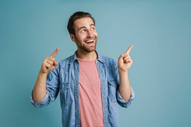 Young bristle man wearing shirt smiling and pointing fingers aside isolated over blue background