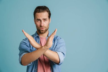 Tişört giyen genç adam mavi arka planda izole edilmiş kameraya dur işareti yapıyor.