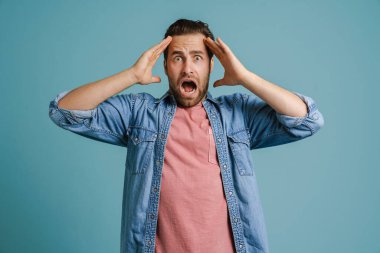 Young man holding his head while expressing surprise isolated over blue background