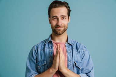 Genç kıllı adam ellerini birarada tutarken gülümsüyor mavi arka planda soyutlanmış.