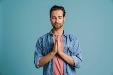 Genç kıllı adam ellerini birarada tutarken meditasyon yapıyor. Mavi arka planda izole edilmiş.