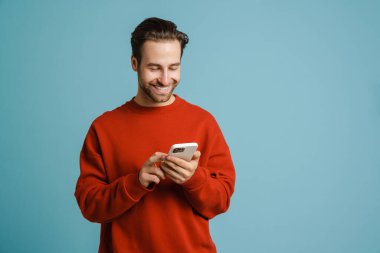 Gülümseyen ve cep telefonu kullanan genç kıllı adam mavi arka planda izole edilmiş.