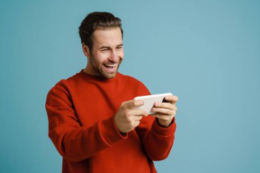 Heyecanlı genç adam gülümsüyor ve mavi arka planda izole bir şekilde cep telefonuyla online oyun oynuyor.