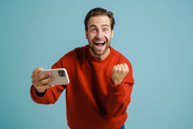 Genç adam başarılı bir jest yapıyor ve mavi arka planda izole edilmiş cep telefonuyla selfie çekiyor.