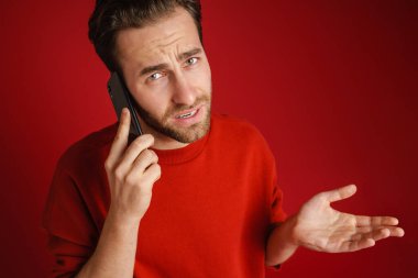 Young bristle man holding copyspace while talking on cellphone isolated over red background