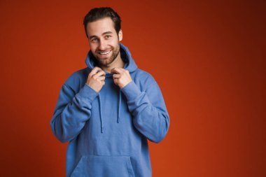 Kapüşonlu genç adam gülümsüyor ve kırmızı arka planda izole edilmiş kameraya bakıyor.