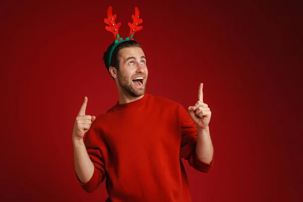 Young Man Wearing Christmas Antlers Pointing Fingers Upward Isolated Red — Φωτογραφία Αρχείου