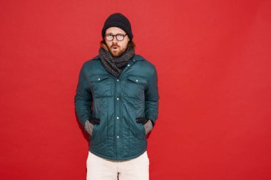 Şapkalı ve ceketli kızıl adam titriyor ve kırmızı arka planda soğuktan soyutlanmış hissediyor.