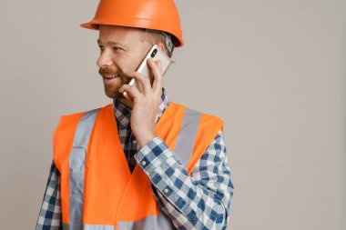 White man worker wearing helmet talking on mobile phone isolated over grey background