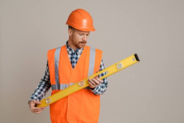 White man worker wearing helmet and vest posing with spirit level tool isolated over grey background
