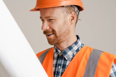 White man worker wearing helmet and vest posing with drawing isolated over grey background