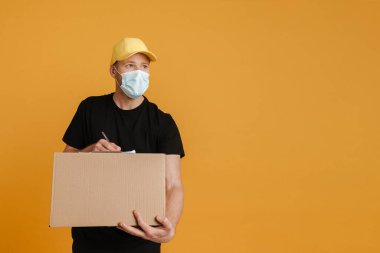 White delivery man in face mask writing down notes on clipboard isolated over yellow background