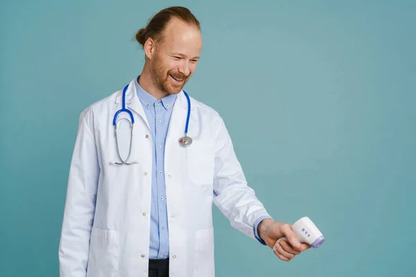 White male doctor wearing lab coat smiling and using thermometer isolated over blue background