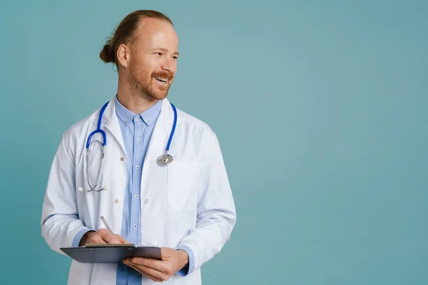 White male doctor wearing lab coat smiling while writing on clipboard isolated over blue background