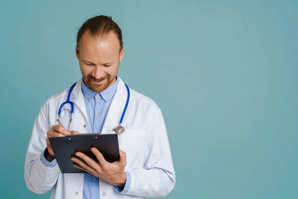 White male doctor wearing lab coat smiling while writing on clipboard isolated over blue background