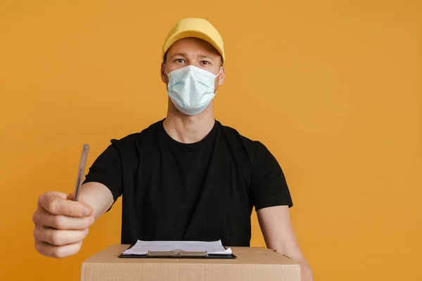 White delivery man in face mask posing with cardboard box and clipboard isolated over yellow background