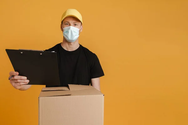 White delivery man in face mask posing with cardboard box and clipboard isolated over yellow background