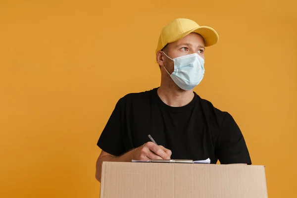 White delivery man in face mask writing down notes on clipboard isolated over yellow background