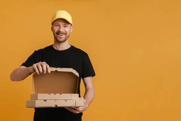 White delivery man in cap smiling while posing with pizza cardboard boxes isolated over yellow background
