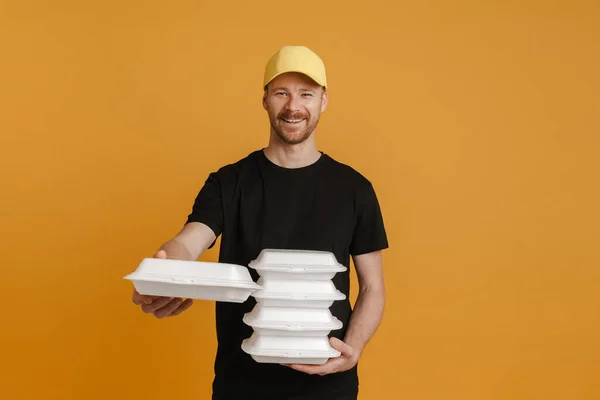 White delivery man in cap smiling while posing with lunch boxes isolated over yellow background