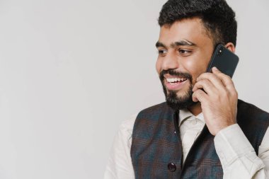 Bearded south asian man wearing vest talking on cellphone isolated over white wall